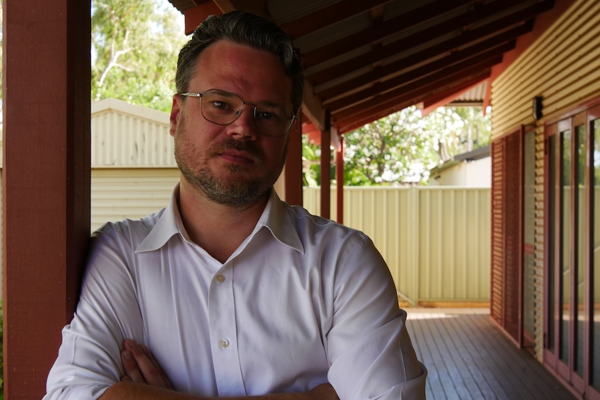 A man in a white shirt leaning against a wooden beam with his arms crossed looking straight ahead.