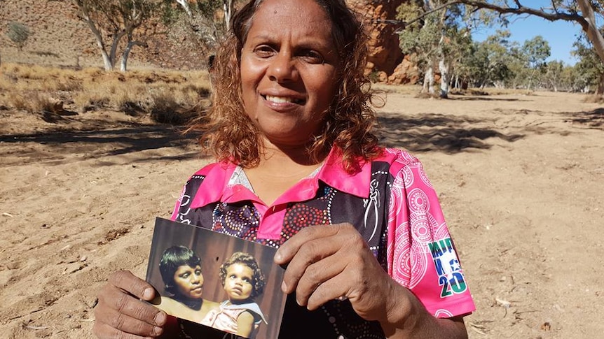Indigenous woman stands smiling in the sunshine surrounding by Outback vegetation and holds old photograph.