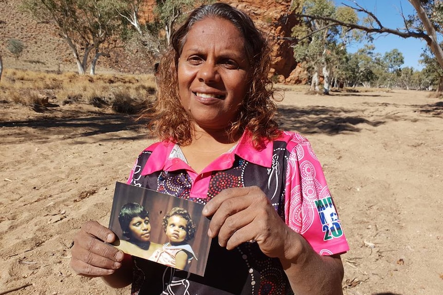 Indigenous woman stands smiling in the sunshine surrounding by Outback vegetation and holds old photograph.