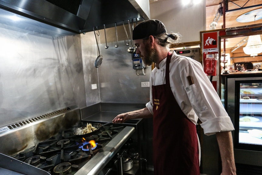 A chef standing at a stove holding a frying pan over a flame.