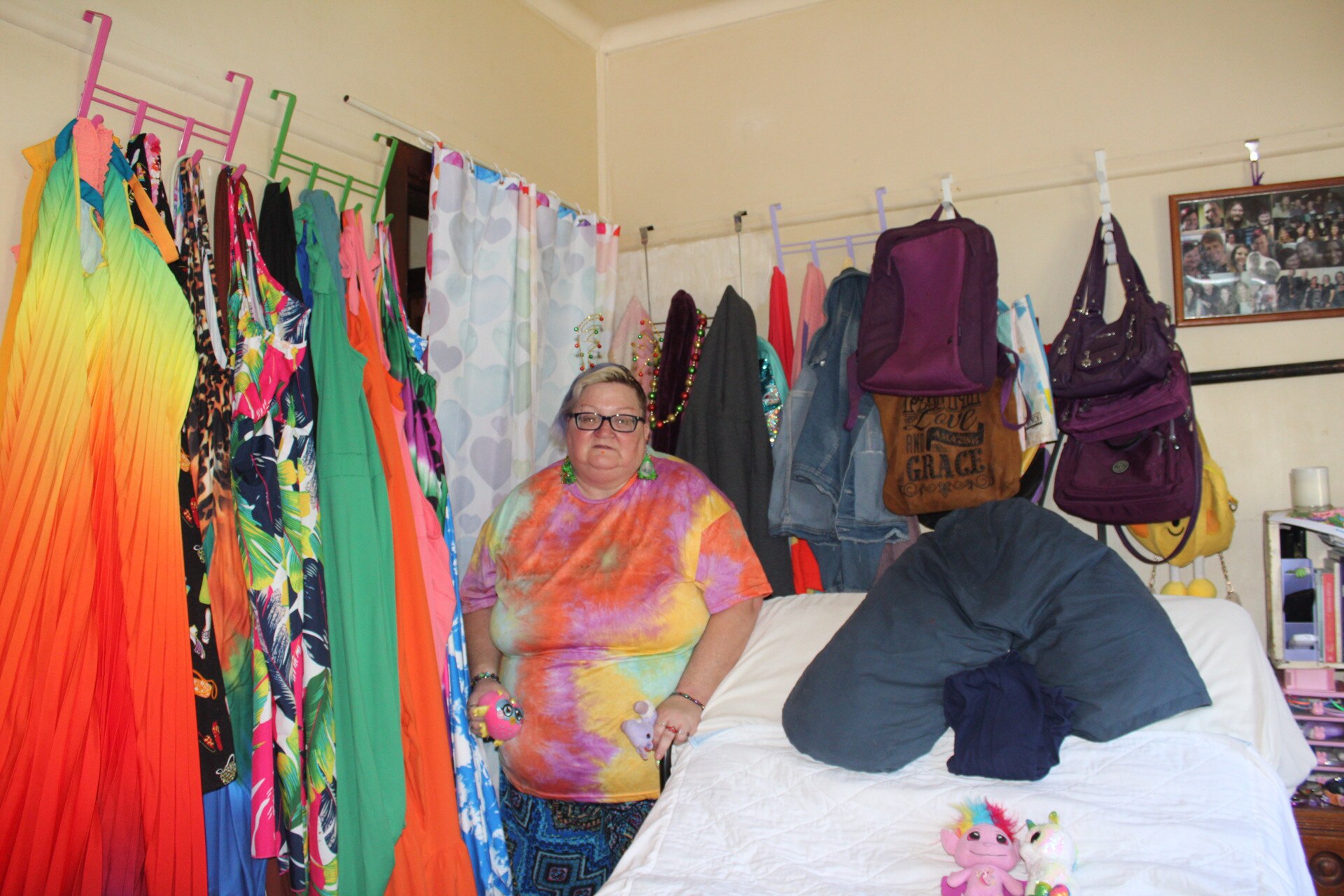 A woman standing next to a bed in a colourful bedroom with lots of coloured clothes hanging from the wall.