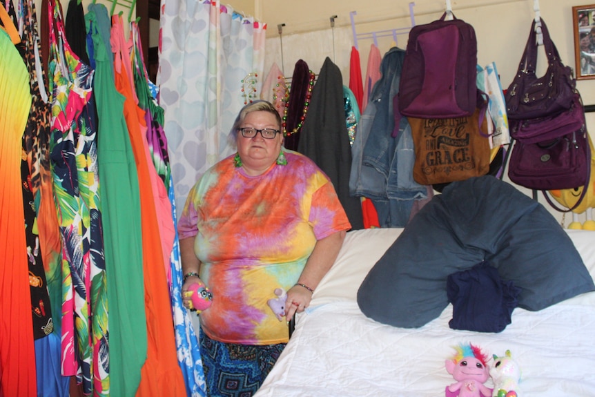 A woman standing next to a bed in a colourful bedroom with lots of coloured clothes hanging from the wall.
