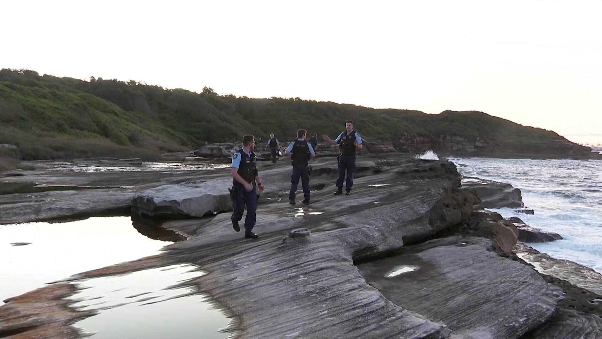 Three police men standing on a rocky shore speak to each other.