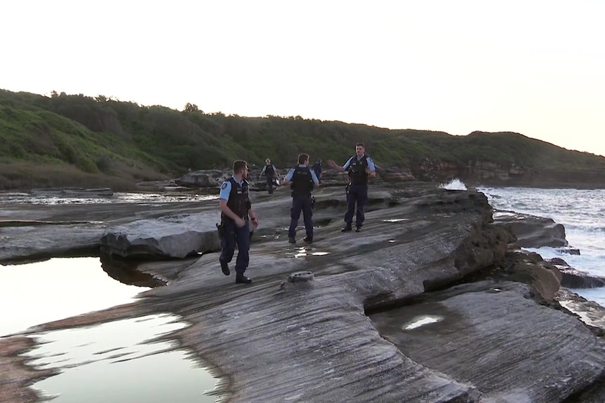 Three police men standing on a rocky shore speak to each other.