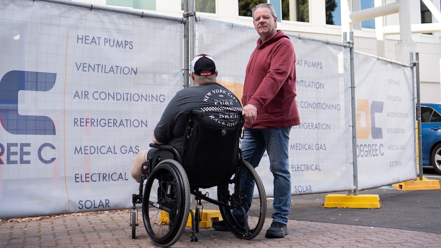A man in a wheelchair is photographed with his back to the camera, while a tall man in a red hoodie stands next to him.