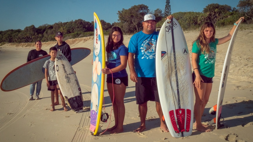 Family portrait on a beach with mother, father and four children holding their surfboards.