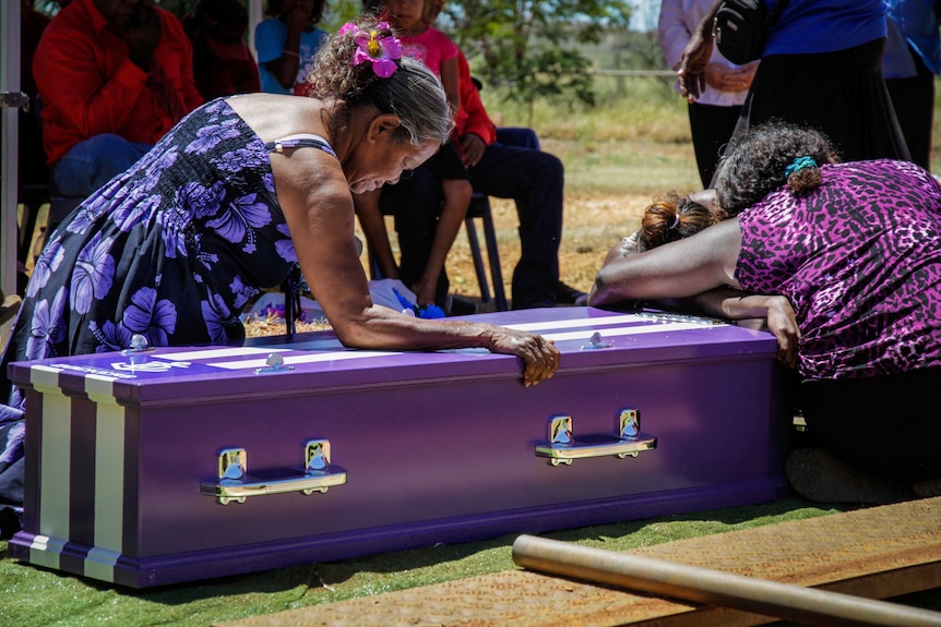 Indigenous women leaning on a purple coffin, crying.