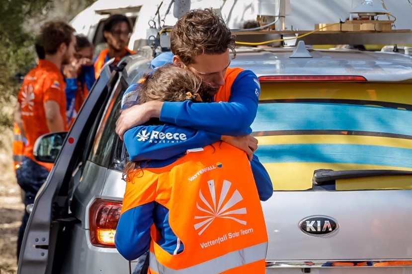 Two people wearing blue long sleeve tops and an orange jacket hug in front of a silver car