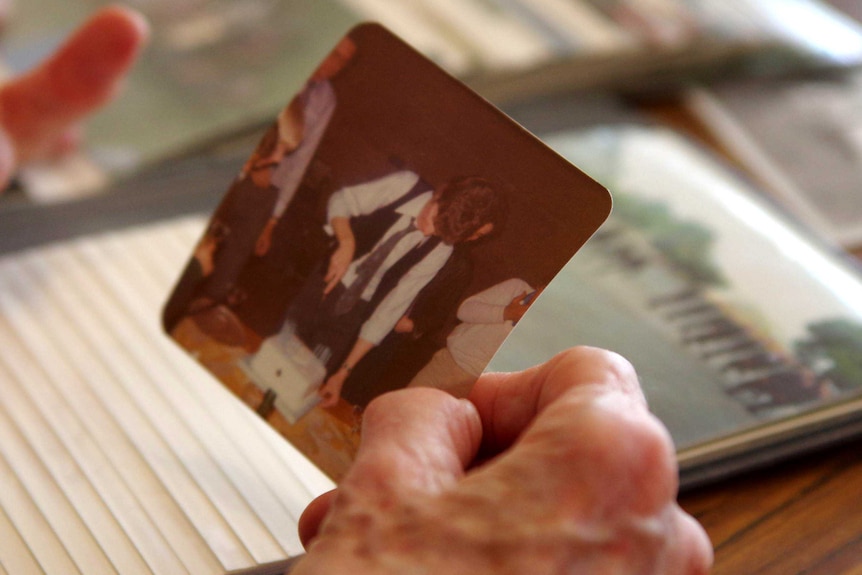 An elderly woman's hand holds a photograph of a man cutting a birthday cake.