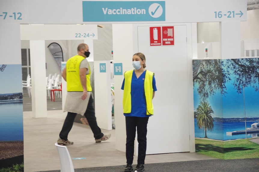 A man and a woman wearing masks and high-vis vests in a large vaccination clinic.