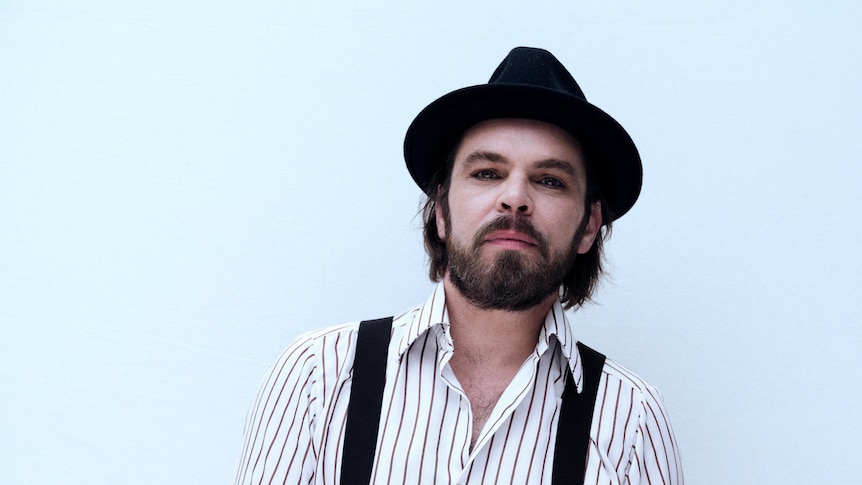 Gaz Coombes wears a striped shirt with suspenders, a fedora hat, and a short beard