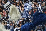 A crowd of Geelong Cats fans in a stadium grandstand, wearing blue and white, holding banners and shaking oversized pom poms.