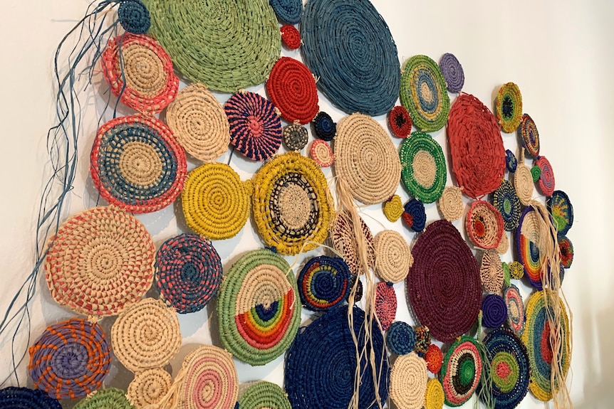 About fifty coloured woven circles sewn together and hang on a wall.