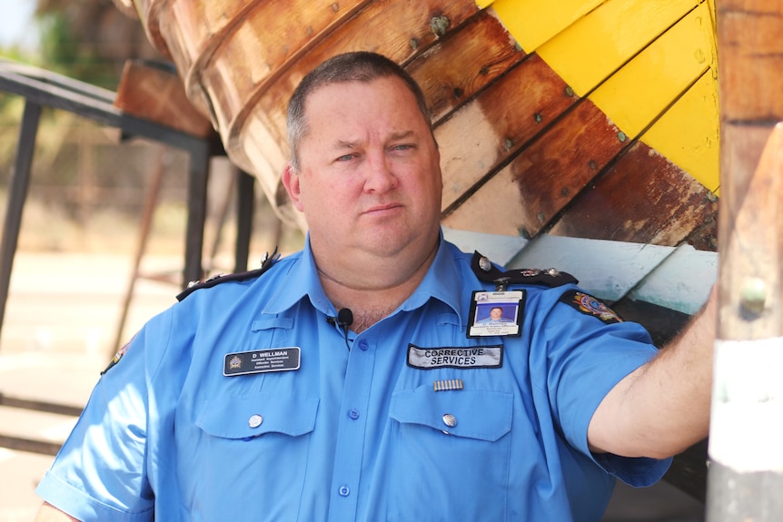 Man in blue prison officer uniform stands under the hull of a wooden boat.