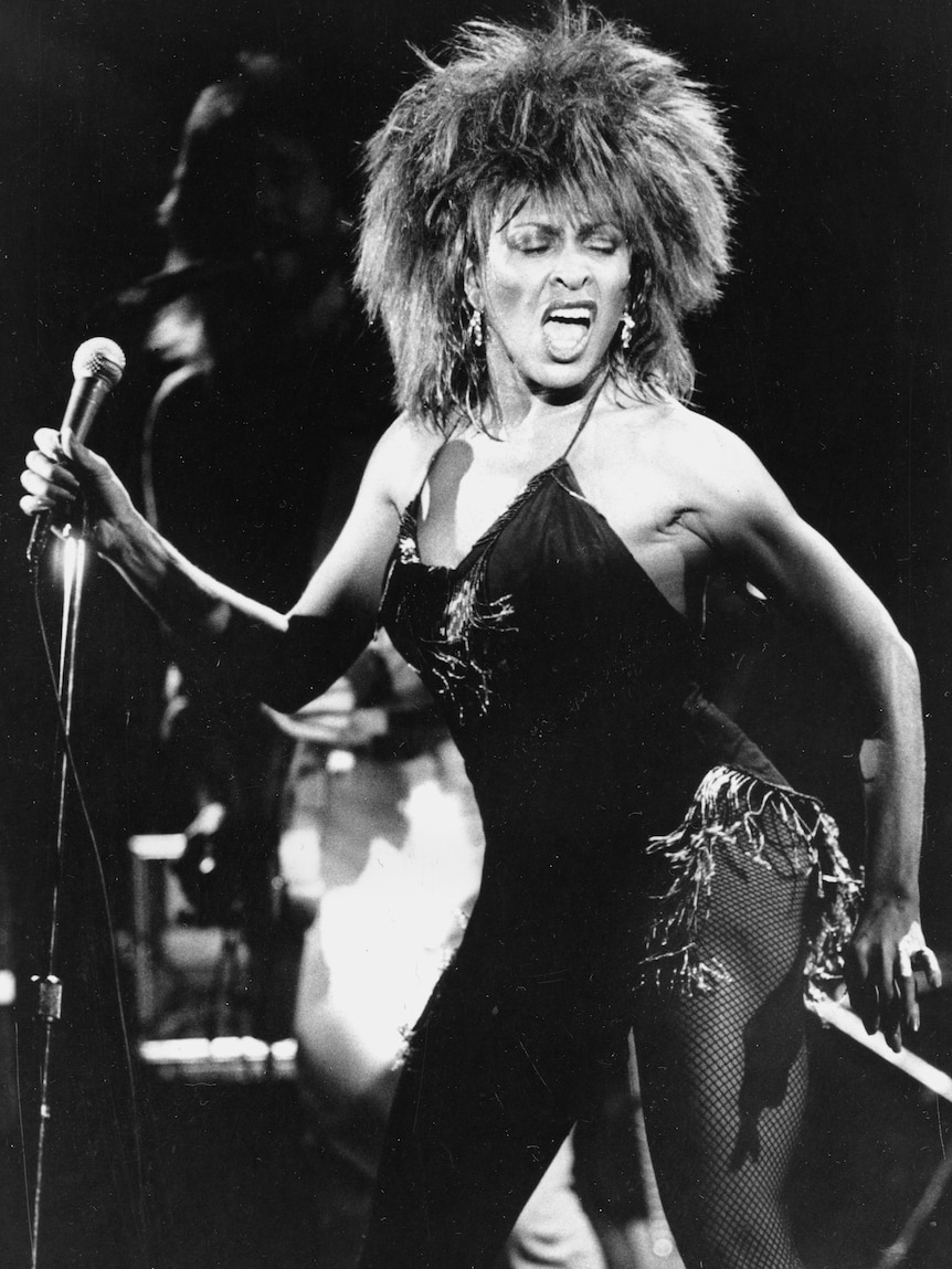 A black-and-white photo of a younger Tina Turner performing on stage wearing fishnet stockings and a short black dress.