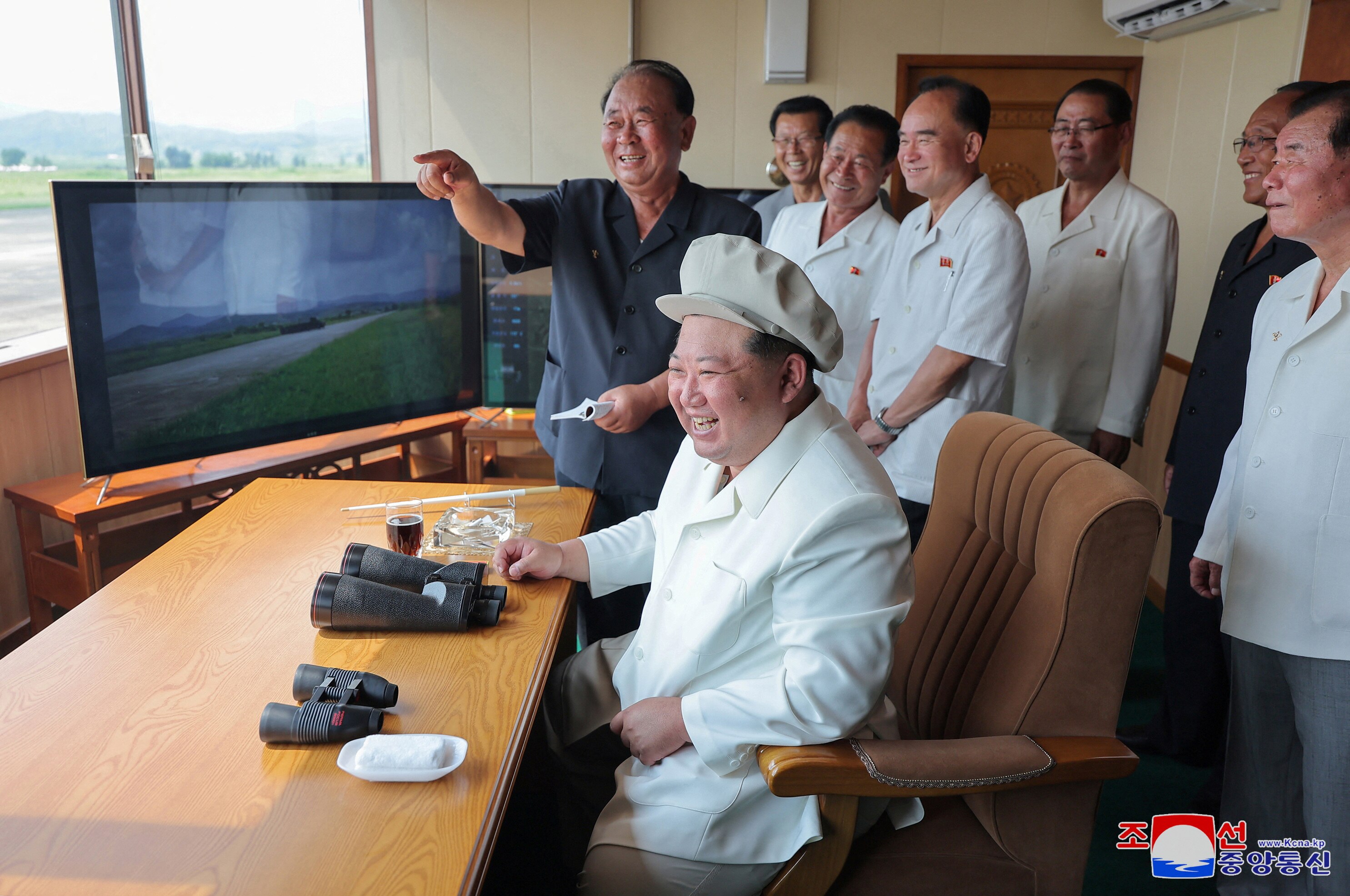 Kim Jong Un wears all white as he smiles and sits at a desk, overseeing a drone test