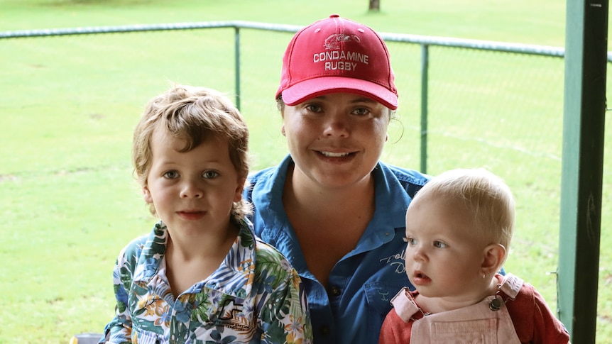 Woman holding toddler and baby on her lap, sitting outdoors and smiling. 