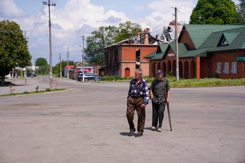 Two men walk through a street, one using a walking stick. The roof of a building behind them has been destroyed.