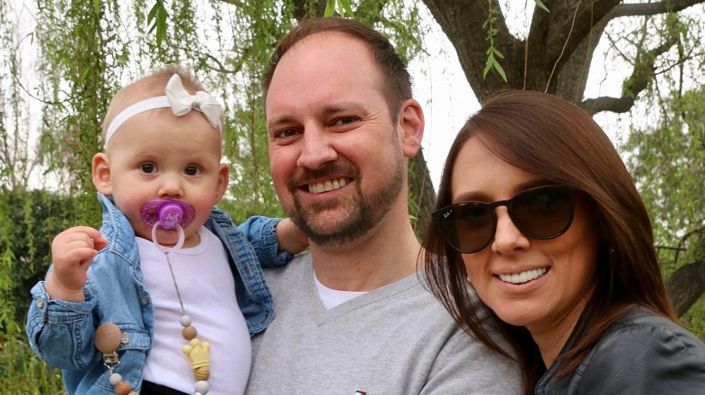 Baby Milana with father Matthew Williams and mother Olia Volova in front of a tree in a park.