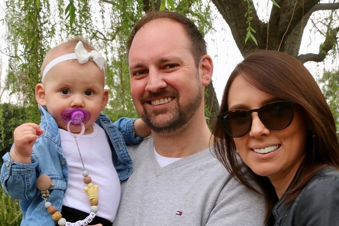 Baby Milana with father Matthew Williams and mother Olia Volova in front of a tree in a park.