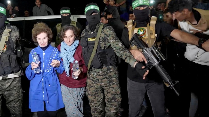 Two women holding water bottles walk next to each other, between men in balaclavas who are holding guns. A crowd behind them