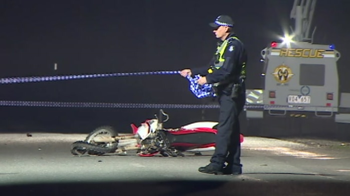 The wreckage of a motorcycle can be seen on the road as a police officer sets up police tape and another speaks on the phone.