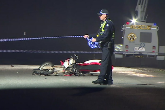 The wreckage of a motorcycle can be seen on the road as a police officer sets up police tape and another speaks on the phone.