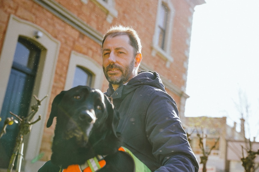 A man crouches down to pat a black labrador dog in front of a Victorian brick building as the sun shines.