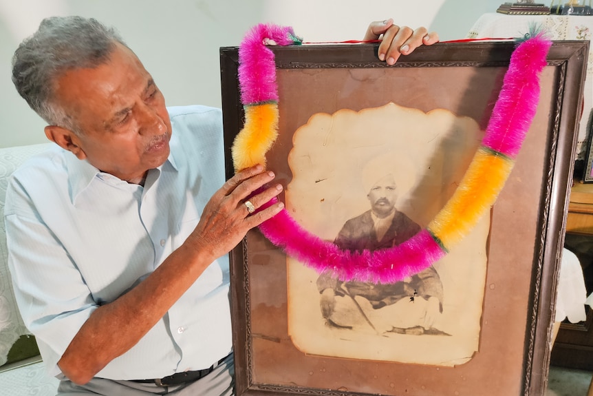 An Indian man stares at a blackand white photograph of his father while holding a pink and yellow ribbon.