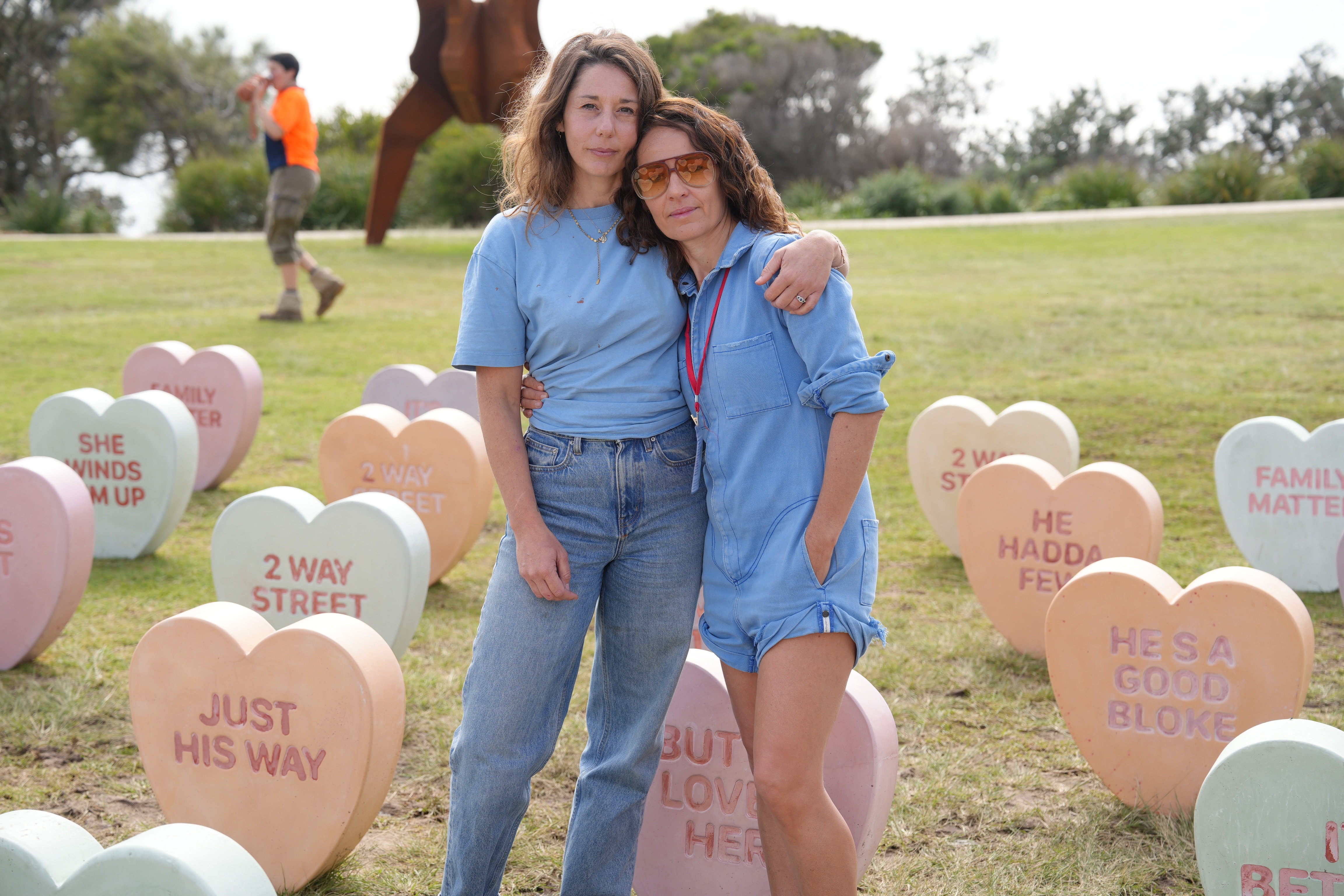 A woman with her arm around another woman standing amongst oversized pastel coloured candy hearts with words on them.