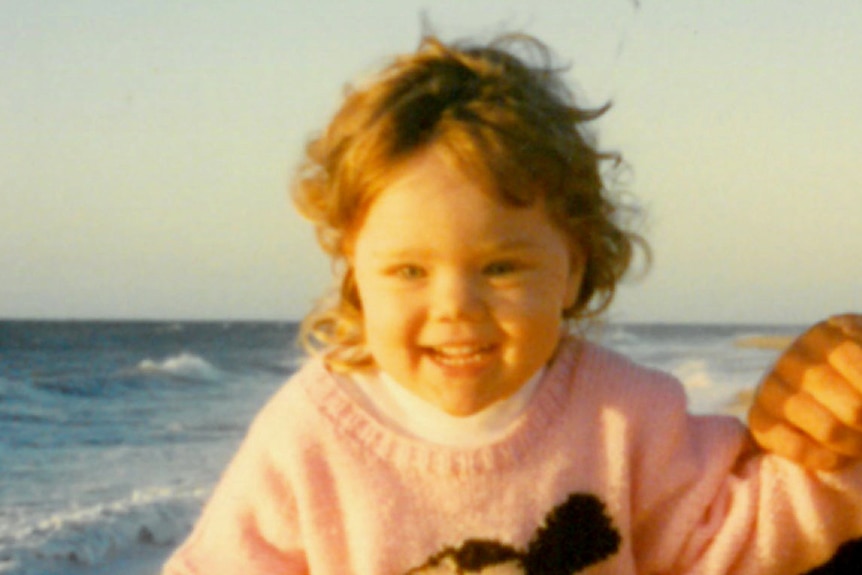 A toddler girl in a knitted jumper with a big smile stands on the beach holding the hand of an adult.