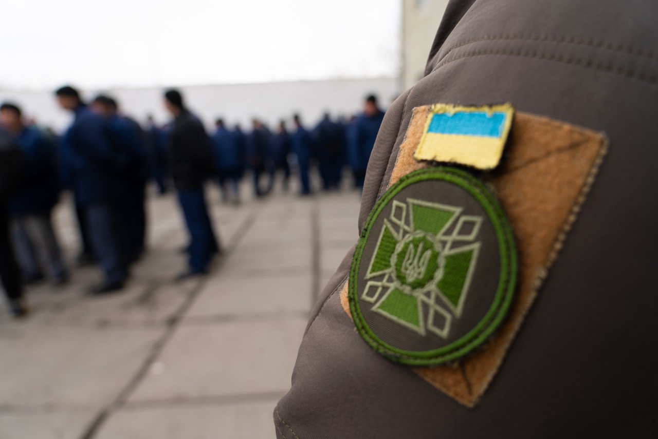 A group of men wearing prison uniforms are visible in the distance behind the arm of a guard wearing a Ukrainian uniform.