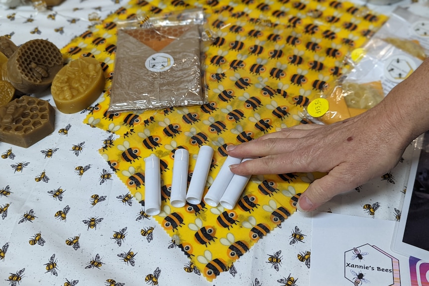 A woman's hand touches bees wax products like a yellow wrap with bees and white tubes.