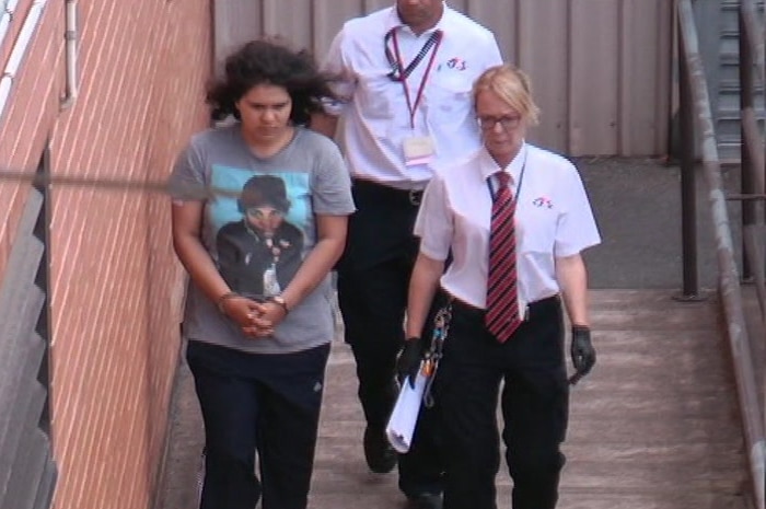 A young woman wearing a t-shirt with a man's face on it walks handcuffed with two court officers
