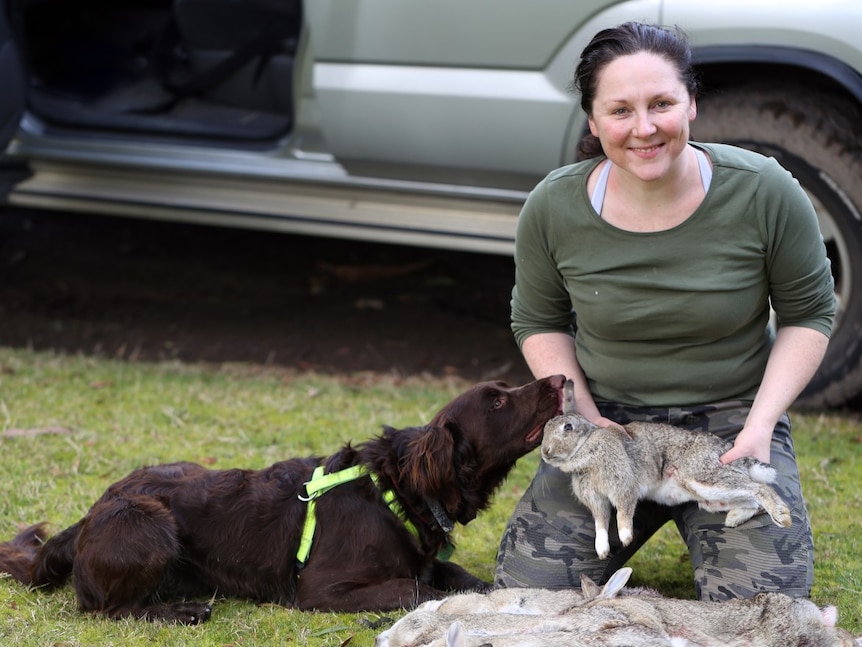 A woman smiling to camera as she holds a dead rabbit, a dog by her side is sniffing her.