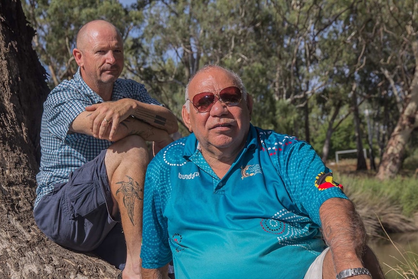 Two men sitting leaning on a tree with a river in the background