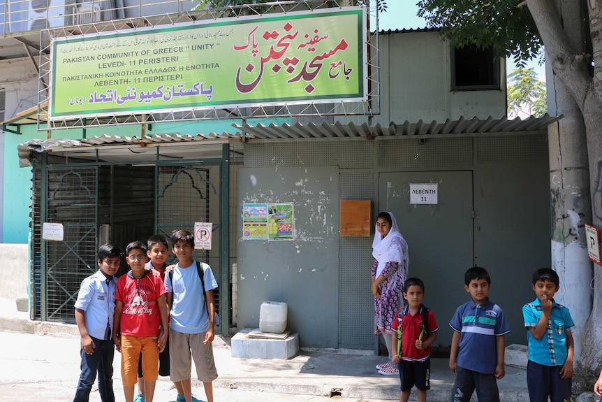 Pakistani children outside an old factory displaying a sign in Greek and Arabic.