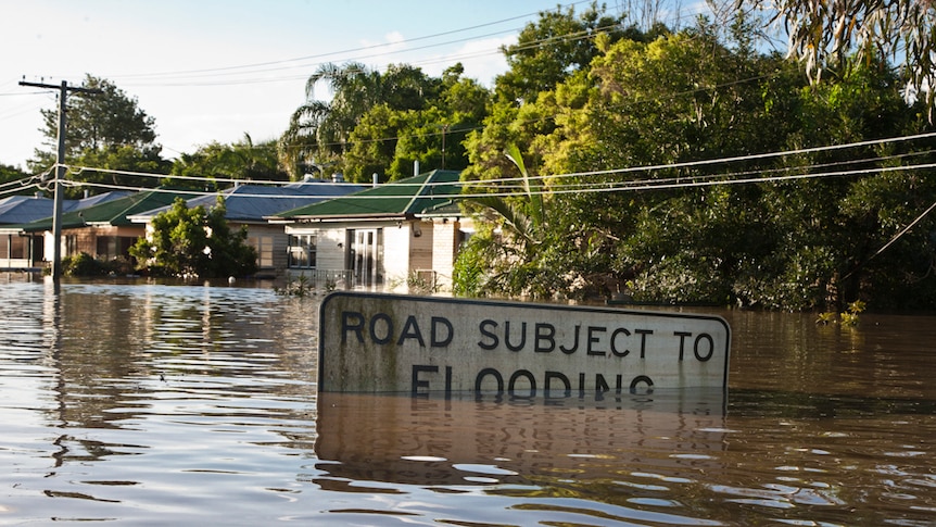 Flood waters almost cover a road sign that says 'Road Subject to Flooding'. Flooded houses in a street in the background.