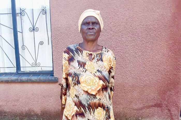 An older woman stands in front of a pink wall.