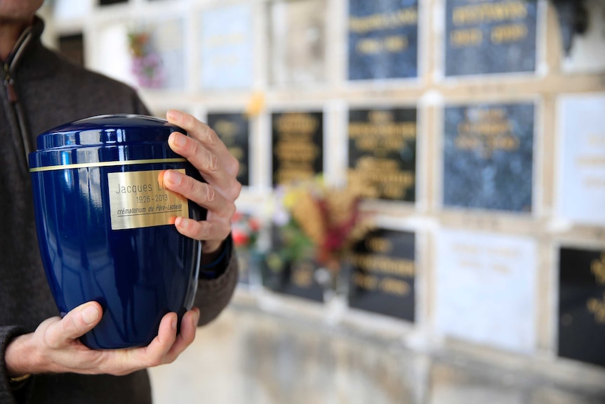 Image of hands holding a blue urn in front of memorial wall.