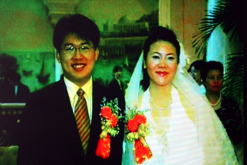 A grainy tinged screenshot shows a happy bride and groom, wearing red flowers on their suit and white dress