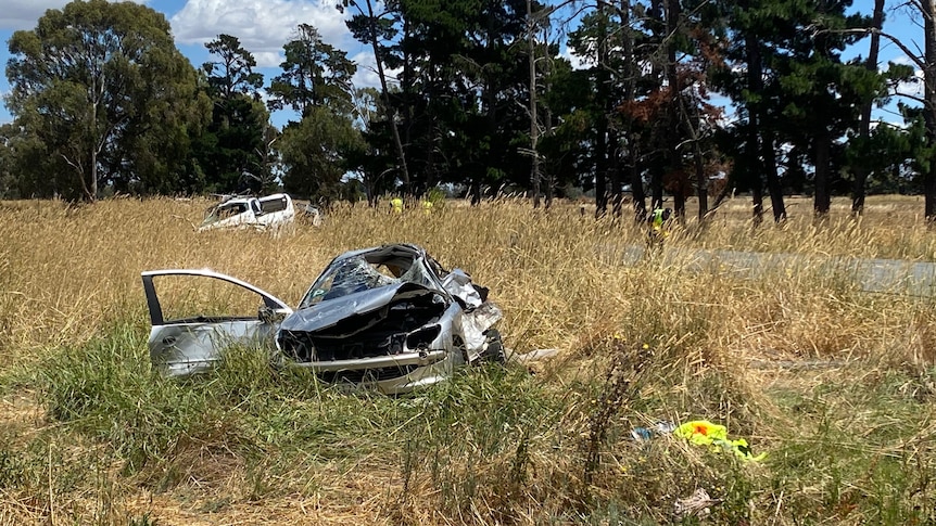 The mangled remains of a silver car lying in a grassy field next a country road.