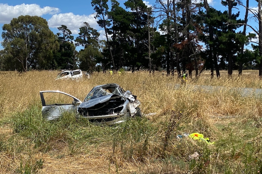 A wrecked silver hatchback in a grassy field. A ute is seen in the background.