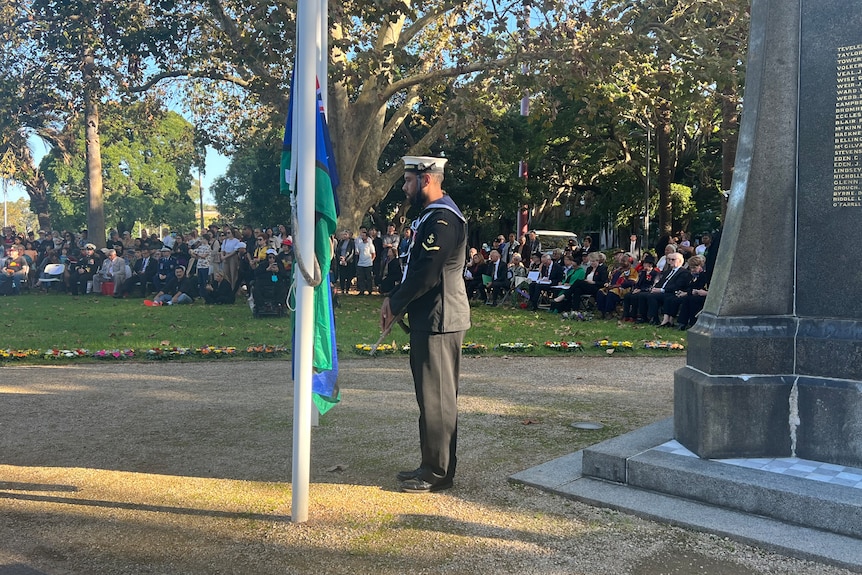 A serviceman stands next to a flagpole while a crowd gathers. 