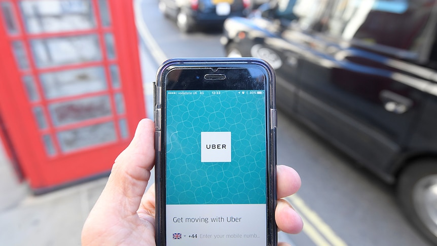 A red telephone box and a black London taxi are seen as a hand holds up a phone displaying the Uber app.