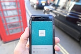 A red telephone box and a black London taxi are seen as a hand holds up a phone displaying the Uber app.