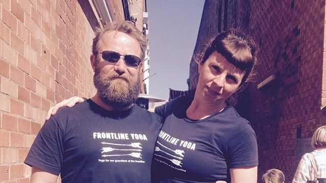 A man and a woman stand with their arms around each other's shoulders wearing black shirts that say Frontline Yoga