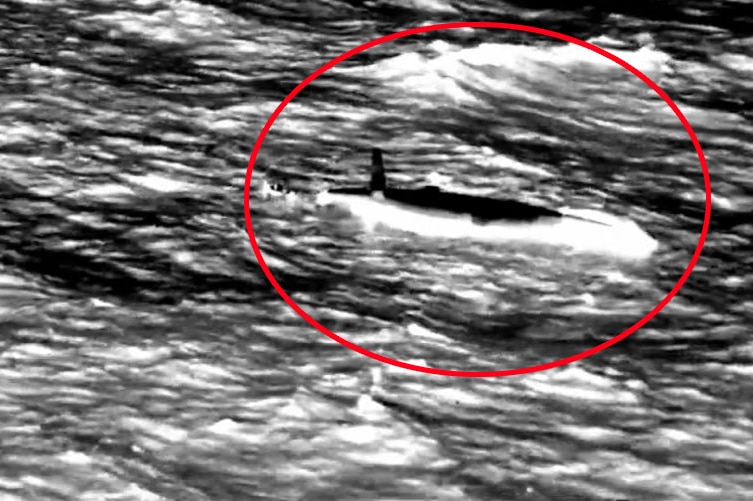 A screenshot of black and white helicopter search footage showing an upturned boat on the water.