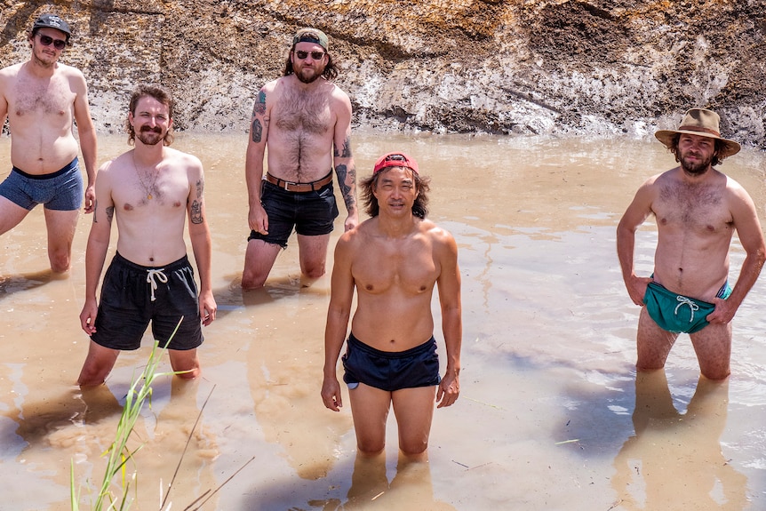 A group of shirtless men stand in a muddy, dam-like hole.