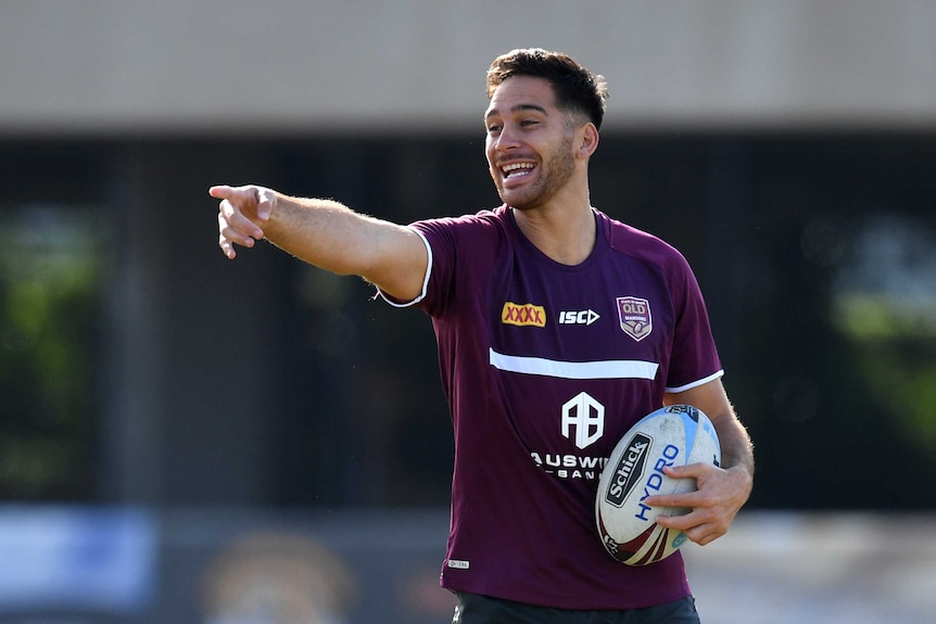 A rugby league male player points a finger on his right hand and holds a ball with his left hand as he laughs.